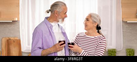 Zufriedenes älteres kaukasisches Paar, das mit Weingläsern in der Küche anstößt und gemeinsam feiert Stockfoto