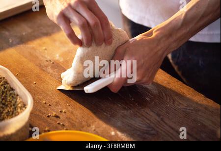 Nahaufnahme von menschlichen Händen, die einen weißen Teigschaber verwenden und rohen, ungebackenen Teig auf einer strukturierten Holzschneideplatte sorgfältig handhaben und formen. Mehl Stockfoto