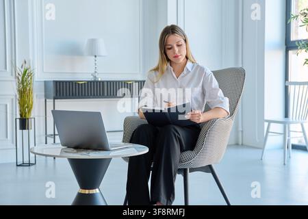Junge weibliche Spezialistin schreibt während der Arbeit in einem modernen Büroraum in ein Notizbuch Stockfoto