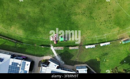 Luftaufnahme zeigt einen Traktor und einen Rasenmäher, die grünes Gras auf einem Sportplatz neben Gebäuden mit Solarpaneelen mähen Stockfoto