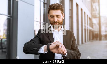 Junger kaukasischer Mann schaut auf seine Smartwatch im Business-Anzug, urbanes Setting. Business-Konzept Stockfoto