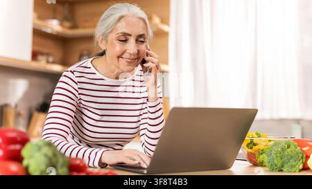 Senior weiß Frau lächelt, während sie Laptop benutzt und am Telefon spricht in der Küche zu Hause Stockfoto