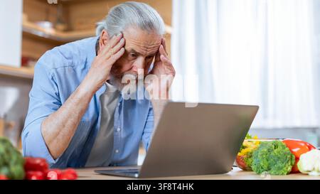 Besorgter älterer kaukasischer Mann, der sich in der Küche über den Computer aufregt, Gewichtsabnahme-Konzept Stockfoto