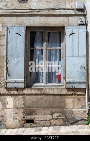 Rustikale, hellblaue Fensterläden in der Rue Jeanne d'Arc in Orléans, Frankreich, umrahmen ein verwittertes Fenster, an dem eine französische Flagge neben den Gardinen erscheint. Stockfoto