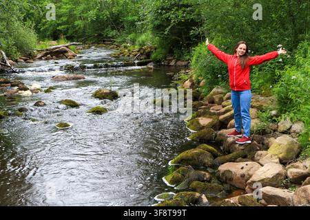 Junge Frau in roter Jacke steht mit ausgestreckten Armen an einem Waldbach Stockfoto