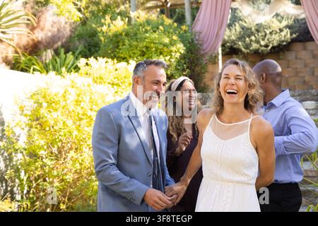 Verschiedene Gruppe Paar hält sich an den Händen und lacht im Garten unter Pergola Frau weißes Kleid Mann grauer Anzug Stockfoto