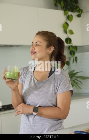 Afroamerikanische Frau hält einen grünen Smoothie und trägt ein graues Sport-T-Shirt an der Küchentheke Stockfoto