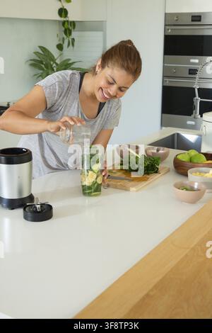 Afroamerikanische Frau gießt Wasser aus einem Glasbehälter in einen Mixbecher mit Bananengrün in der Küche Stockfoto