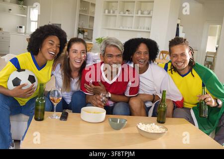 Verschiedene Freunde in Fußballtrikots im Wohnzimmer, die sich vorbeugen und ein Spiel mit Ball und Bier beobachten Stockfoto