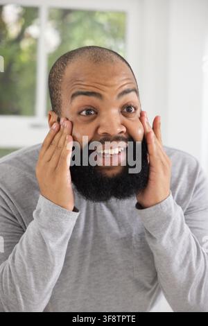 Erwachener Mann drückt Hände an die Wangen, lächelt, Bart sichtbar, trägt graues Hemd an einem hellen Fenster Stockfoto