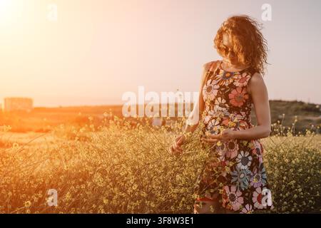 Frau Blumen Sonnenuntergang, lockenköpfige junge Frau in Blumenkleid, die während des warmen goldenen Lichts der Abenddämmerung in einem Feld gelber Wildblumen steht Stockfoto