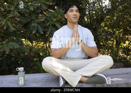 Asiatischer Mann sitzt im Park mit gekreuzten Beinen auf einer Bank in blauem Hemd, Perlenarmband, Flasche links Stockfoto
