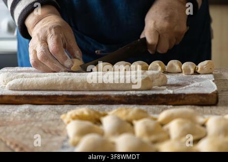 Hände schneiden einen Zylinder frischen Teigs in gleichmäßige Portionen für die Herstellung traditioneller hausgemachter Knödel, wobei ein Prozess sorgfältiger Vorbereitung eingefangen wird für Stockfoto
