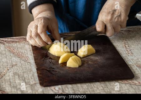 Seniorhände schneiden gekonnt rohe gelbe Kartoffelkeile mit einem scharfen Metallküchenmesser, was kulinarische Aktivität, häusliches Leben und Vorbereitung vermittelt Stockfoto