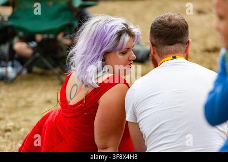 FRAU MIT LILA HAAREN UND EINEM RÜCKEN-TATTOO, REPORTAGE, GREEN MAN FESTIVAL 2016, BRECON, WALES, UK, 2016: Eine junge Frau mit kurzen lila Haaren, Glitzer auf der Wange und einem kreisförmigen Tattoo auf dem Rücken blickt über ihre Schulter, während sie mit einem Mann in einem weißen T-Shirt sitzt und spricht. Foto: Rob Watkins. INFO: Das Green Man Festival begann 2003 in Wales. Jährlich findet es am dritten Wochenende im August in den Brecon Beacons statt und feiert alternative Musik, Kunst, Literatur, Wissenschaft und familienfreundliche Kultur in einer wunderschönen Berglandschaft. Stockfoto