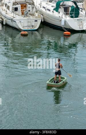 Junger Mann paddelt ein kleines Plastikschlauchboot im Stehen im Yachthafen des Lymington River, Hampshire, UK Stockfoto