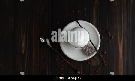Ansicht von oben eines schaumigen Cappuccinos in einer Glas Tasse mit einer Schokoladentorte, einem Löffel und Kaffeebohnen, die auf einem dunklen Holztisch verstreut sind. Stockfoto