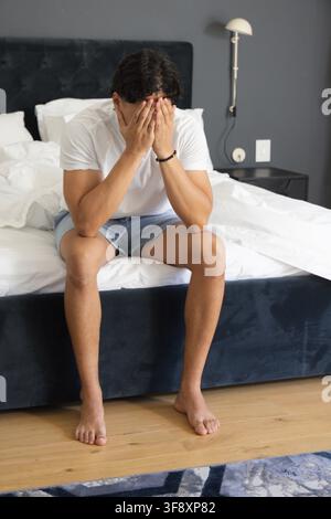Asiatischer erwachsener Mann sitzt auf der Bettkante im Schlafzimmer, lehnt sich zurück, bedeckt das Gesicht und trägt ein schwarzes Perlenarmband Stockfoto
