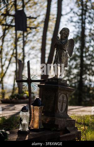 Eine alte Engelsstatue, die auf einem Steingrab mit Gedenklaternen und einem Kreuz auf einem Friedhof betet. Schöner verschwommener Waldhintergrund mit sanftem Sonnenlicht Stockfoto