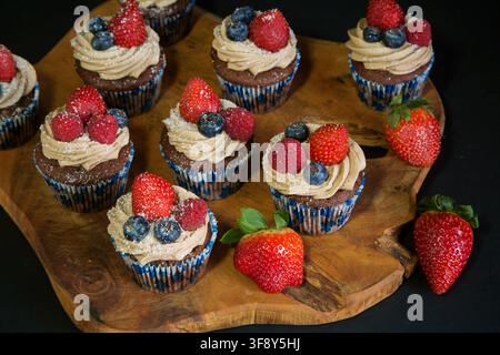 Schokoladen-Cupcakes, die mit Erdbeeren, Blaubeeren und Himbeeren auf einem Holzbrett auf schwarzem Hintergrund dekoriert sind. Hochwertiges Foto Stockfoto