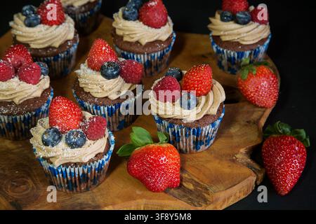 Schokoladen-Cupcakes, die mit Erdbeeren, Blaubeeren und Himbeeren auf einem Holzbrett auf schwarzem Hintergrund dekoriert sind. Hochwertiges Foto Stockfoto
