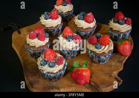 Schokoladen-Cupcakes, die mit Erdbeeren, Blaubeeren und Himbeeren auf einem Holzbrett auf schwarzem Hintergrund dekoriert sind. Hochwertiges Foto Stockfoto