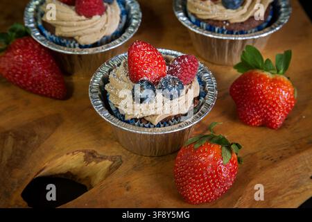 Schokoladen-Cupcakes, die mit Erdbeeren, Blaubeeren und Himbeeren auf einem Holzbrett auf schwarzem Hintergrund dekoriert sind. Hochwertiges Foto Stockfoto