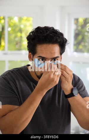 Afroamerikanischer Mann verwendet blaue Gel-Patches an seinem Heimspiegel in der Nähe weißer Fenster, graues T-Shirt, Smartwatch Stockfoto