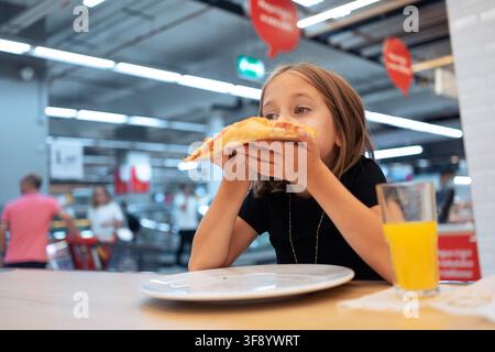 Mädchen isst eine Scheibe Pizza und trinkt Saft, während sie an einem Tisch in einer Foodcourt in einem Einkaufszentrum sitzt. Stockfoto
