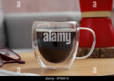 Frisch gebrühter schwarzer Kaffee in einer klaren doppelwandigen Glastasse mit einer roten Moka-Kanne im Hintergrund, eine rote Moka-Kanne ist im verschwommenen Hintergrund zu sehen Stockfoto