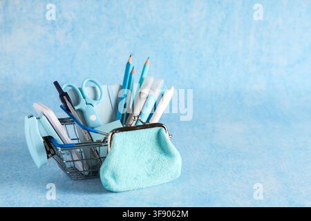 Einkaufswagen für die Schulanfangseinkäufe mit blauem Schreibwaren und Münztäschchen auf hellblauem Hintergrund Stockfoto