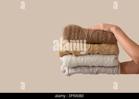 Frauenhand hält einen ordentlich gefalteten Stapel warmer Kabelstrickpullover in neutralen Farben, isoliert auf einem beigen Hintergrund Stockfoto