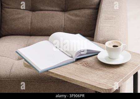 Offenes Notizbuch mit Millpapier und Stift neben einer Tasse Kaffee auf einem Holzbeistelltisch neben einem Sofa, Neben einem bequemen Sofa, eine entspannte Arbeit oder ein entspanntes Lernen Stockfoto