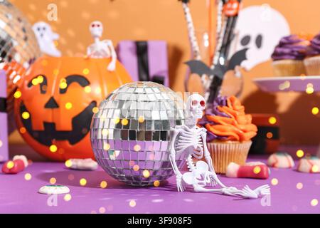 Halloween-Party-Dekorationen mit einem Skelett, einem Discokugel, einem Kürbis und Cupcakes auf einem lila und orangefarbenen Hintergrund Stockfoto