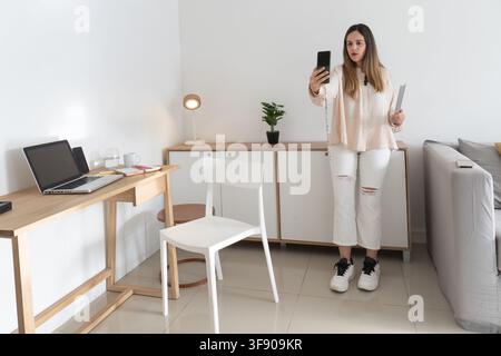 Vollständige Aufnahme einer Frau, die ihr Handy hochhält, während sie sich selbst filmt, steht in einem gefliesten Wohnzimmer in der Nähe eines Schreibtisches, einer Anrichte und eines Sofas, lässiges Outfit Stockfoto
