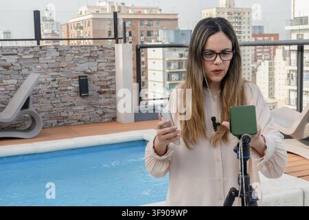 Nahaufnahme einer Frau mit Brille, die ein Smartphone in der Nähe eines Stativaufbaus hält, dichte Stadtlandschaft und Dachpool im Hintergrund sichtbar Stockfoto