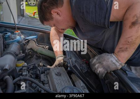 Mechaniker in einem dunklen T-Shirt und Schutzhandschuhen, der während einer DIY-Reparatur im Freien den Motorraum eines Autos inspiziert und arbeitet Stockfoto