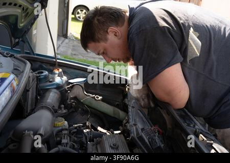 Mechaniker in einem dunklen T-Shirt und Schutzhandschuhen, der während einer DIY-Reparatur im Freien den Motorraum eines Autos inspiziert und arbeitet Stockfoto