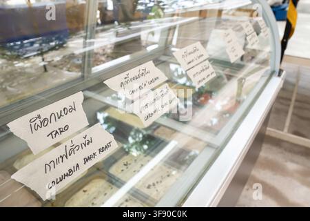 Gläserner Eisschank mit handgeschriebenen thailändischen Etiketten und englischen Übersetzungen für verschiedene Geschmacksrichtungen wie Taro, Kaffee, Vanille, Joghurt, Passionsfrucht Stockfoto