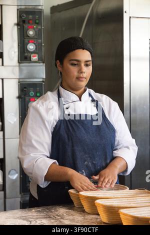 Afroamerikanische Frau trägt Chefjacke, blaue Schürze und arbeitet an einer Backbank mit Bannetons Stockfoto