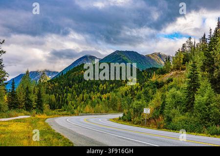Eine kurvenreiche zweispurige Asphaltstraße, die durch einen dichten Wald aus immergrünen und herbstfarbenen Bäumen unter majestätischen alaskischen Bergen führt. Stockfoto