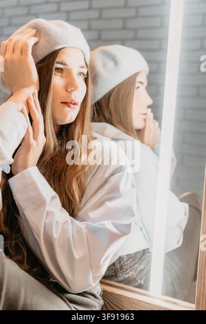 Frau mit Barett spiegelt junge Dame in weißem Hemd, die ihren Hut berührt, während sie in einen Studiomirror schaut Stockfoto