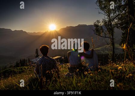 Drei Freunde, die sich vor einem atemberaubenden Sonnenuntergang über sanften Bergen abzeichnen. Sie sitzen auf einem grasbewachsenen Hügel, haben die Arme umeinander gelegt und genießen th Stockfoto