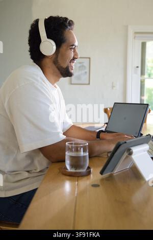 Afroamerikaner Mitte 30, der an einem Esstisch-Arbeitsplatz sitzt und Laptop, Tablet, Kopfhörer verwendet Stockfoto