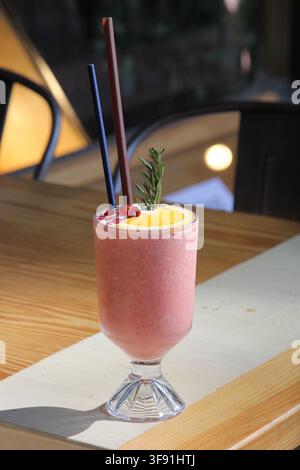 Ein erfrischender rosa Frucht-Smoothie, serviert in einem hohen Glas mit einem Strohhalm und einer Obstgarnitur auf einem Holztisch. Stockfoto