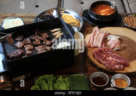 Korea-Barbecue-Fest mit Schweinebauch, auf einer Pfanne gegrilltem Rindfleisch, Beilagen, Suppe und Salat auf einem Tisch. Stockfoto