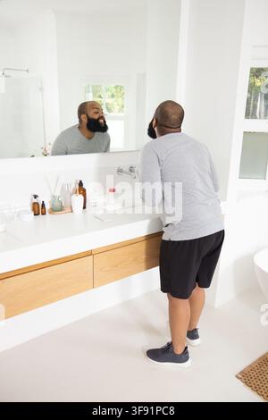 Afroamerikaner inspiziert Bart am weißen Waschbecken, Spiegel im Badezimmer in grauem Hemd, schwarzen Shorts Stockfoto