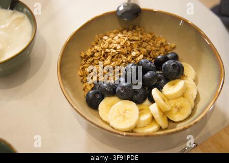 Keramik-Frühstücksschüssel auf hellem Tisch in der Küche mit Granola, Blaubeeren, Banane Stockfoto