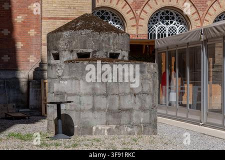 Eine verwitterte betonartige Bunkerstruktur steht auf Kies in einem Hof neben einem Backsteingebäude mit gewölbten Fenstern. Stockfoto