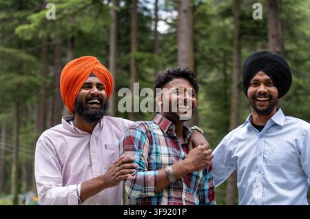 Bustbild von drei männlichen Freunden, darunter zwei junge Sikh-Männer, die zusammen stehen und herzlich lachen, während sie vor einem Kiefernwald posieren. Stockfoto